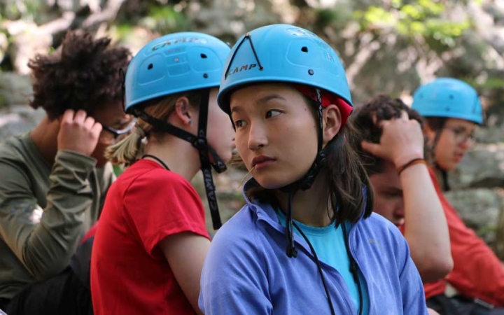 A group of young people appear to prepare to rock climb. A few of them are wearing helmets. 